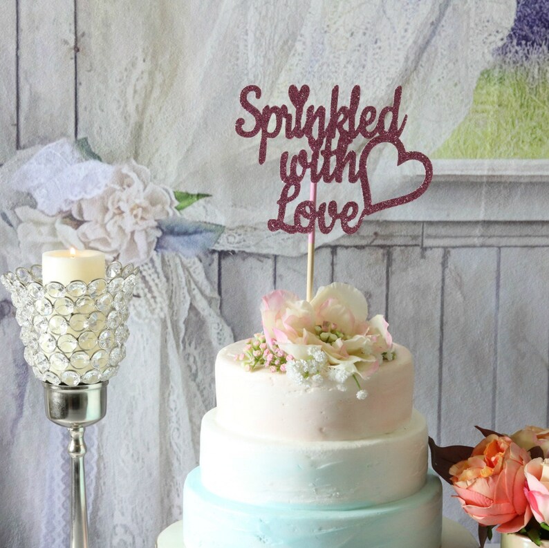 May include: A three-tiered wedding cake decorated with a pink and blue ombre frosting. The cake is topped with a pink glitter cake topper that says "Sprinkled with Love" with a heart.