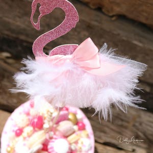 K&ouml;nnte beinhalten: Rosa glitzernder Flamingo-Kuchentopper mit einem wei&szlig;en Feder- und T&uuml;llrock und einer rosa Satin Schleife. Der Topper befindet sich auf einem Kuchen, der mit rosa und goldenen S&uuml;&szlig;igkeiten verziert ist.