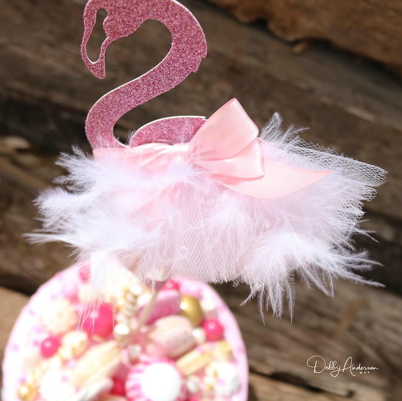 K&ouml;nnte beinhalten: Rosa glitzernder Flamingo-Kuchentopper mit einem wei&szlig;en Feder- und T&uuml;llrock und einer rosa Satin Schleife. Der Topper befindet sich auf einem Kuchen, der mit rosa und goldenen S&uuml;&szlig;igkeiten verziert ist.