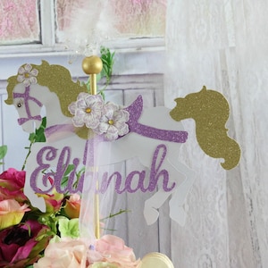 May include: White glitter carousel horse cake topper with a purple glitter mane and tail. The horse has a purple ribbon and flowers on its head. The name "Eliana" is written in purple glitter on the horse's body.