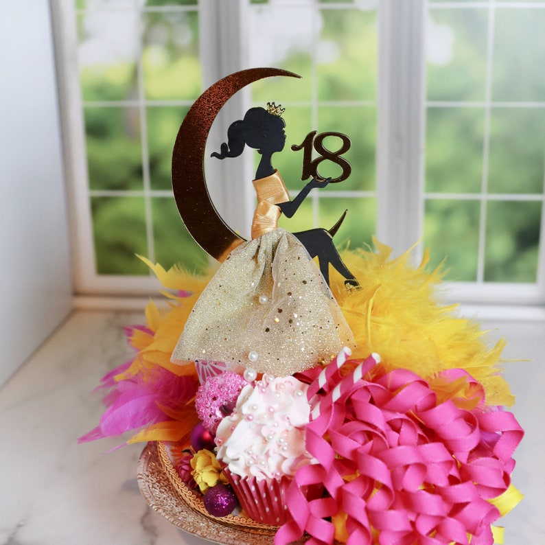 Peut inclure: Une d&eacute;coration de g&acirc;teau repr&eacute;sentant la silhouette noire d'une personne couronn&eacute;e assise sur un croissant de lune, tenant le nombre 18. La figurine porte une jupe dor&eacute;e scintillante, entour&eacute;e de plumes jaunes et de rubans roses.