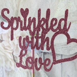 May include: Pink glitter cake topper with the words "Sprinkled with Love" and a heart shape.