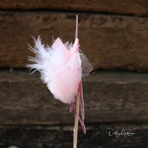 K&ouml;nnte beinhalten: Ein rosa und wei&szlig;er Cupcake-Topper aus Feder und T&uuml;ll mit einem rosa Glitzerstern. Der Topper ist an einem Holzstiel befestigt.