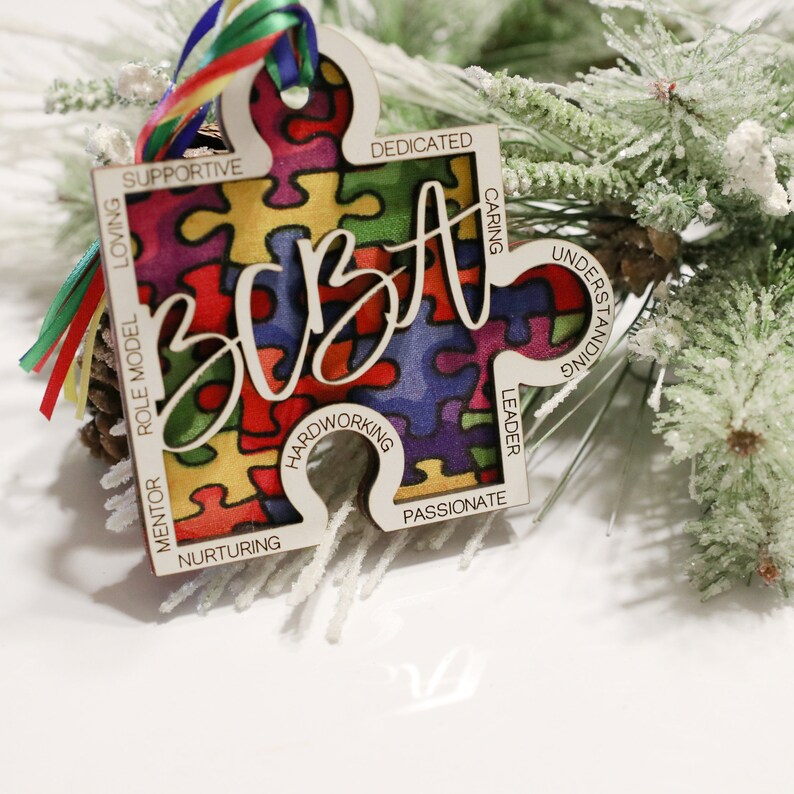 May include: A wooden puzzle piece ornament with a colourful puzzle pattern and the word "BEST" written in cursive. The ornament has words around the edge, including "supportive", "loving", "role model", "mentor", "nurturing", "hardworking", "passionate", "leader", "caring", "understanding", and "dedicated".