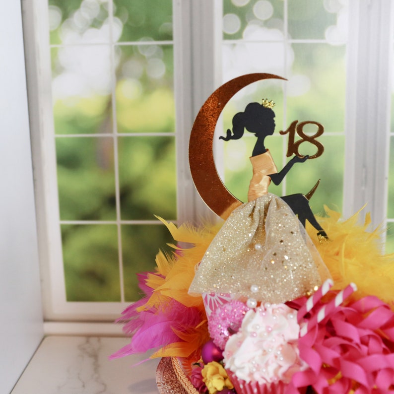 Peut inclure: Une d&eacute;coration de g&acirc;teau repr&eacute;sentant la silhouette noire d'un personnage assis sur un croissant de lune dor&eacute; m&eacute;tallique, tenant le nombre 18 en or. Le personnage porte une jupe dor&eacute;e scintillante avec de petites perles blanches et des plumes.