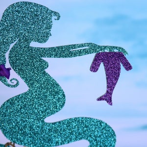 May include: A teal glitter mermaid silhouette holding a purple glitter mermaid silhouette. The mermaid is pregnant and has a bow on her tail.