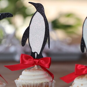 Pu&ograve; includere: Tre cupcake decorati con topper a forma di pinguino nero e fiocchi rossi. I pinguini sono fatti di carta glitterata e hanno la pancia bianca. I cupcake sono glassati con glassa bianca e hanno una spolverata di granella.
