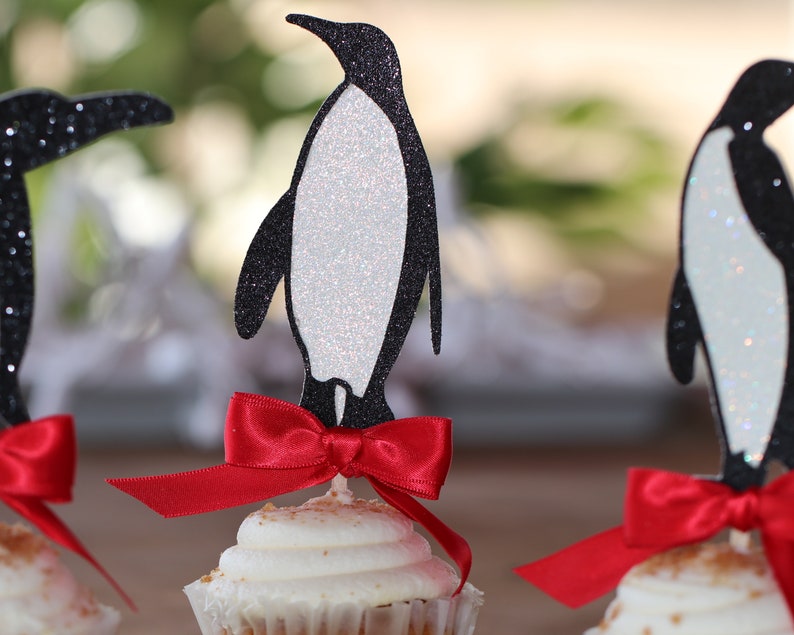 Pu&ograve; includere: Tre cupcake decorati con topper a forma di pinguino nero e fiocchi rossi. I pinguini sono fatti di carta glitterata e hanno la pancia bianca. I cupcake sono glassati con glassa bianca e hanno una spolverata di granella.