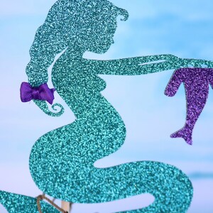 May include: A teal glitter mermaid silhouette with a purple bow on her hair, holding a purple glitter dolphin.