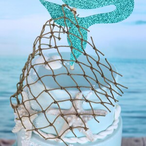 May include: A two-tiered cake decorated with a blue mermaid tail, brown fishing net, and white starfish. The cake is on a blue and white background.