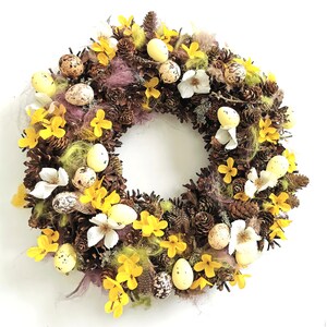 Puede incluir: Una corona de primavera con piñas marrones, flores amarillas y huevos de Pascua moteados.
