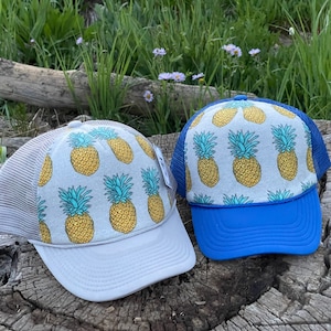 Op de afbeelding: Twee trucker caps met ananasprint. De ene pet is grijs met een wit mesh achterpaneel en een grijze klep. De andere pet is grijs met een blauw mesh achterpaneel en een blauwe klep.