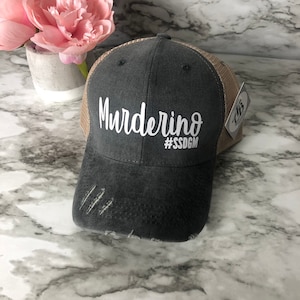 Peut inclure: Un chapeau de camionneur gris et marron avec le texte "Murderino #SSDG" en blanc. Le chapeau a un aspect vieilli avec quelques déchirures dans le tissu.