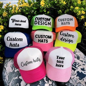 May include: A collection of trucker hats with different colors and designs. The hats are all white with a mesh back and feature various text and designs, including "Custom Hats", "Custom Design", and "Your text here".