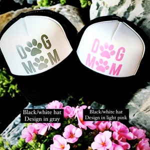 May include: Two black and white trucker hats with a white mesh back. The hats have the text "DOG MOM" in grey and light pink on the front. The hats are sitting on a bed of pink flowers.
