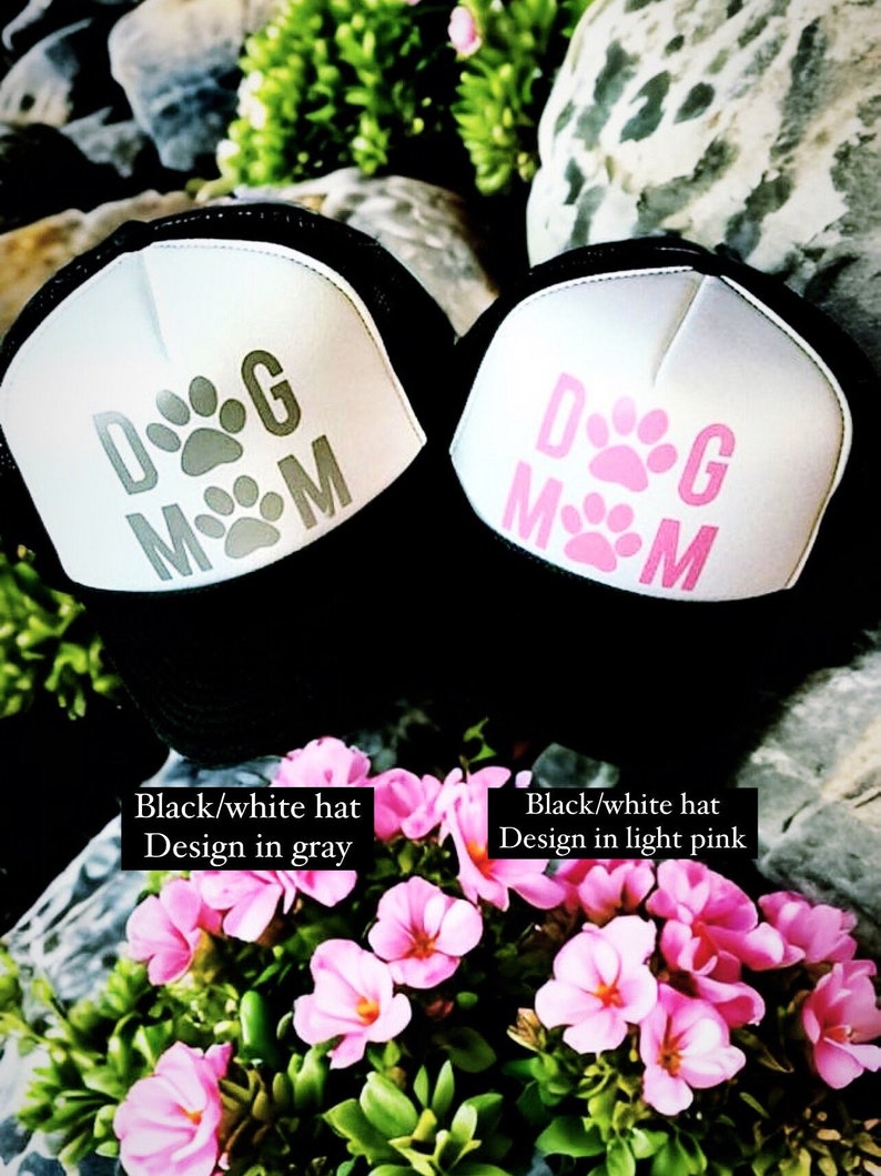 May include: Two black and white trucker hats with a white mesh back. The hats have the text "DOG MOM" in grey and light pink on the front. The hats are sitting on a bed of pink flowers.