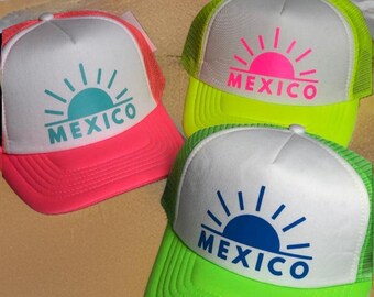 May include: Three neon trucker hats with a white crown and a mesh back. Each hat features a white sun with a different coloured centre and the word "MEXICO" underneath. The hats are on a sandy surface.