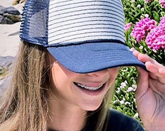 Könnte beinhalten: Eine junge Person trägt eine blaue und weiße gestreifte Trucker-Kappe mit Netzrücken. Die Kappe hat ein marineblaues Schild. Die Person lächelt und trägt eine Zahnspange.
