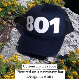 May include: A navy blue trucker hat with a white mesh back. The hat has a white 801 printed on the front. The hat is pictured on a rock with yellow flowers.