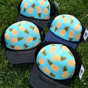 Op de afbeelding: Drie zwarte en blauwe mesh trucker caps met een turquoise kroon met een ananaspatroon. Het ananaspatroon is geel en bruin op een turquoise achtergrond.