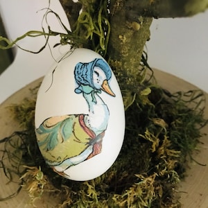Könnte beinhalten: Ein weißes Ei mit einer gemalten Illustration einer Gans mit blauer Haube und grüner und gelber Weste. Das Ei wird auf Moos und einer Holzscheibe präsentiert, mit einem Ast im Hintergrund.