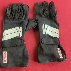 Puede incluir: Un par de guantes de carreras negros con el nombre de la marca "SIMPSON" en un rect&aacute;ngulo rojo en la mu&ntilde;eca. Cada guante tiene un logotipo "S" blanco y una franja blanca en el dorso de la mano.
