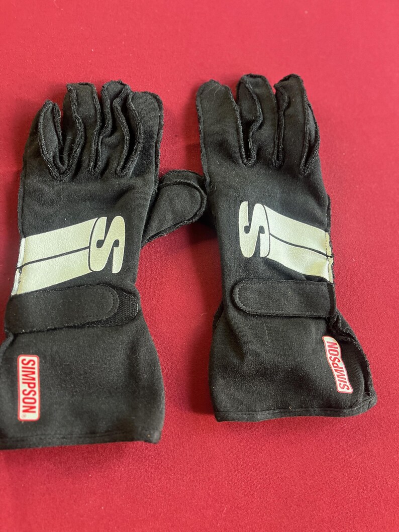 Puede incluir: Un par de guantes de carreras negros con el nombre de la marca "SIMPSON" en un rect&aacute;ngulo rojo en la mu&ntilde;eca. Cada guante tiene un logotipo "S" blanco y una franja blanca en el dorso de la mano.