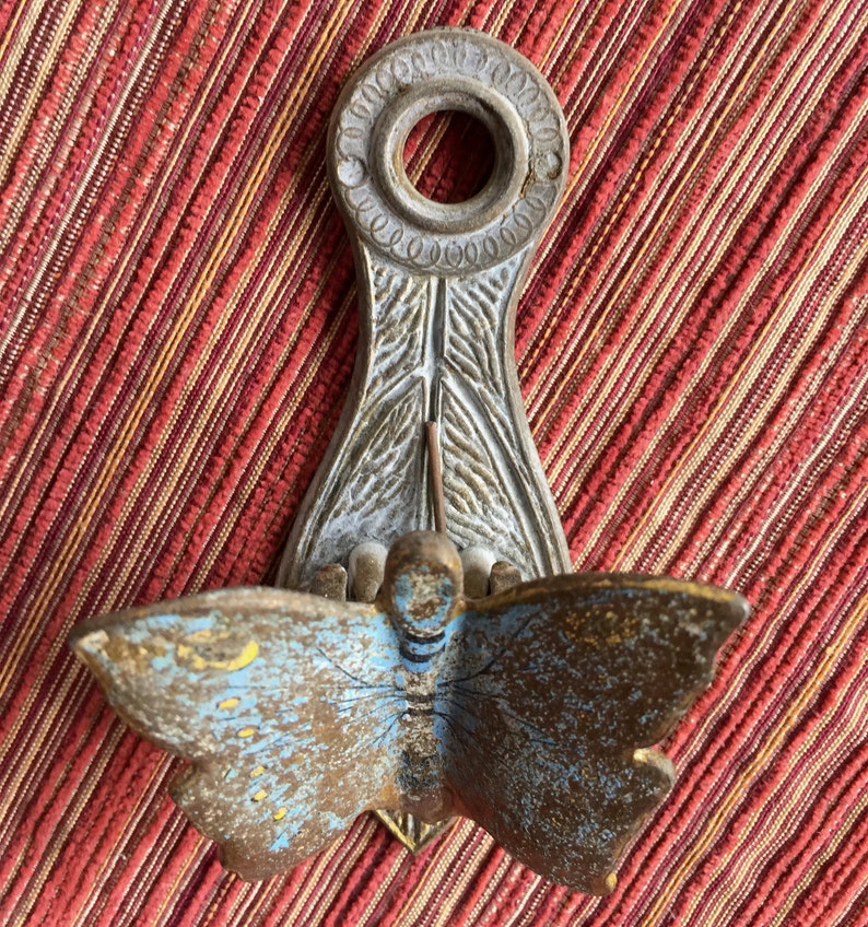 May include: An antique metal wall sconce with a butterfly design. The butterfly has blue and brown wings with yellow accents. The sconce has a decorative top with a circular opening and a detailed, leaf-like base.