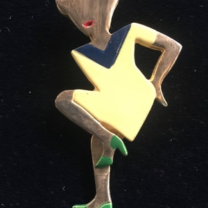 Puede incluir: Un broche dorado que representa a una mujer estilizada con un vestido amarillo y un corpi&ntilde;o azul y negro. La mujer lleva zapatos verdes y tiene la boca roja.
