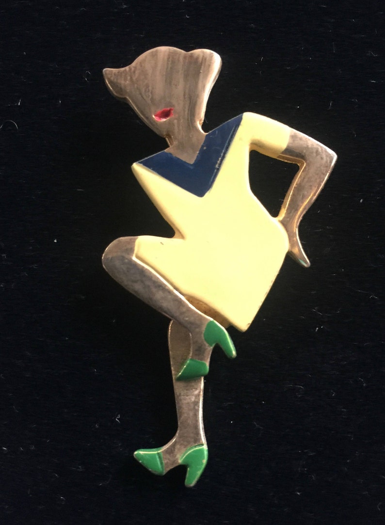 Puede incluir: Un broche dorado que representa a una mujer estilizada con un vestido amarillo y un corpi&ntilde;o azul y negro. La mujer lleva zapatos verdes y tiene la boca roja.