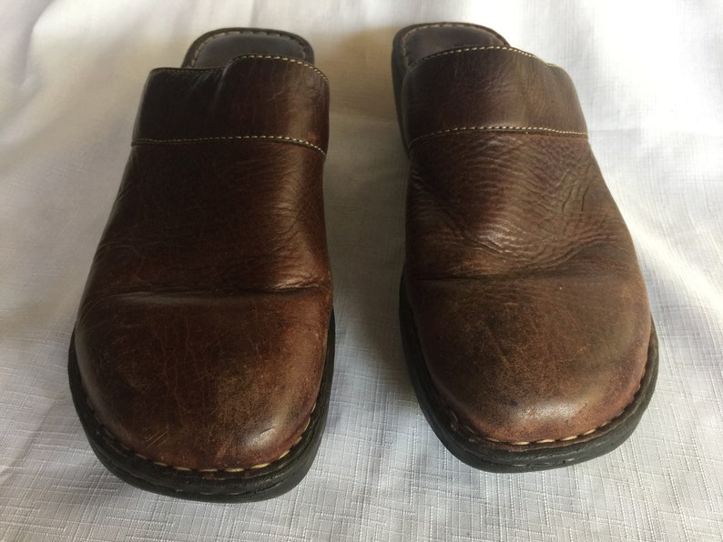 May include: A pair of brown leather slip-on clogs with a black rubber sole.