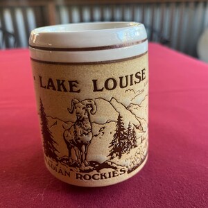 May include: A cylindrical ceramic shot glass with a beige background and brown accents. The glass features an illustration of a ram, mountains, and trees, with the words "LAKE LOUISE" and "CANADIAN ROCKIES" printed on it.