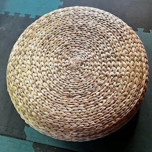 Puede incluir: Un cojín redondo tejido con materiales naturales, de color marrón claro y con un patrón circular.