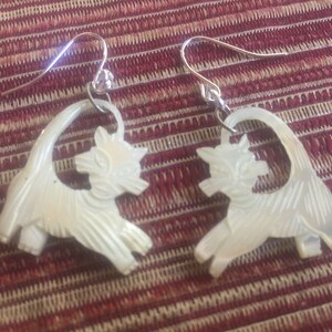 May include: A pair of white mother-of-pearl earrings shaped like cats. The earrings are dangle style with silver hooks.