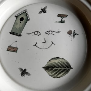 May include: White ceramic bowl with a whimsical design featuring a smiling face, a birdhouse, bees, a leaf, and a sign that says "Welcome".
