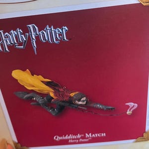 Może przedstawiać: Ozdoba Harry Potter Quidditch Match przedstawiająca postać w bordowo-złotej szacie lecącą na miotle, sięgającą po złoty znicz z różowymi skrzydłami. Bordowe tło zawiera logo Harry Potter oraz napis Quidditch Match.