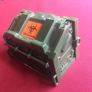 May include: A green plastic box with a biohazard symbol and the word "BIOHAZARD" printed on the top. The box has a hinged lid and a latch.