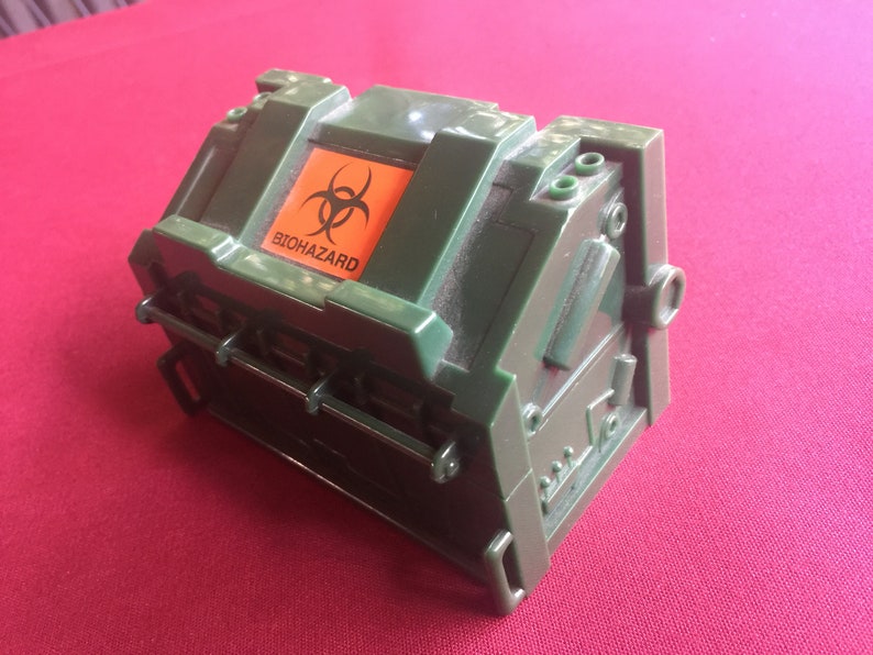 May include: A green plastic box with a biohazard symbol and the word "BIOHAZARD" printed on the top. The box has a hinged lid and a latch.