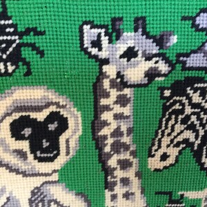 May include: A close-up of a colorful needlepoint tapestry featuring a giraffe, a monkey, and a zebra. The animals are stitched in shades of white, black, and gray against a green background.