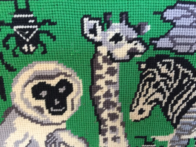 May include: A close-up of a colorful needlepoint tapestry featuring a giraffe, a monkey, and a zebra. The animals are stitched in shades of white, black, and gray against a green background.