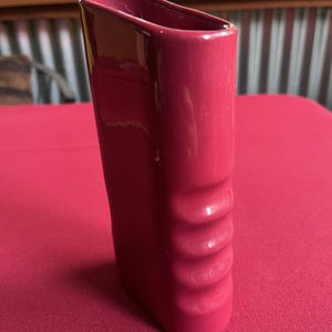 May include: A tall, rectangular, magenta-colored ceramic vase. The vase has a smooth, glossy finish with a series of curved indentations on one side. The vase is set on a magenta surface.