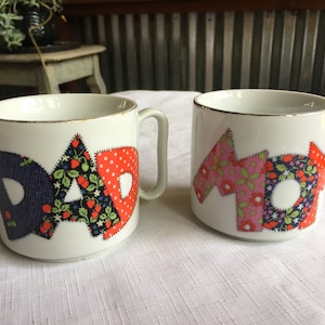 May include: Two white ceramic mugs with gold trim. The mugs have a patchwork design with the words "DAD" and "MOM" spelled out in different colored fabrics. The fabrics have floral and polka dot patterns.