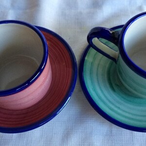 May include: Two ceramic teacups and saucers. The saucer on the left has a pink and red design with a blue rim. The saucer on the right has a green and blue design with a blue rim.
