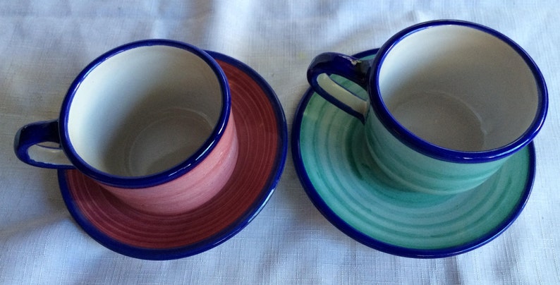 May include: Two ceramic teacups and saucers. The saucer on the left has a pink and red design with a blue rim. The saucer on the right has a green and blue design with a blue rim.