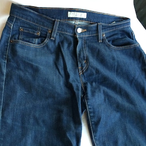 Puede incluir: Un par de pantalones vaqueros de mezclilla azul oscuro con un estilo clásico de cinco bolsillos. Los vaqueros tienen una pierna recta y un lavado ligeramente descolorido.