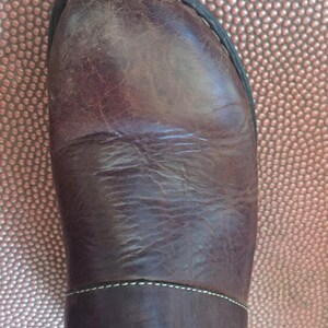 May include: Close-up of the top of a brown leather boot with a white stitch detail. The boot is on a brown textured surface.