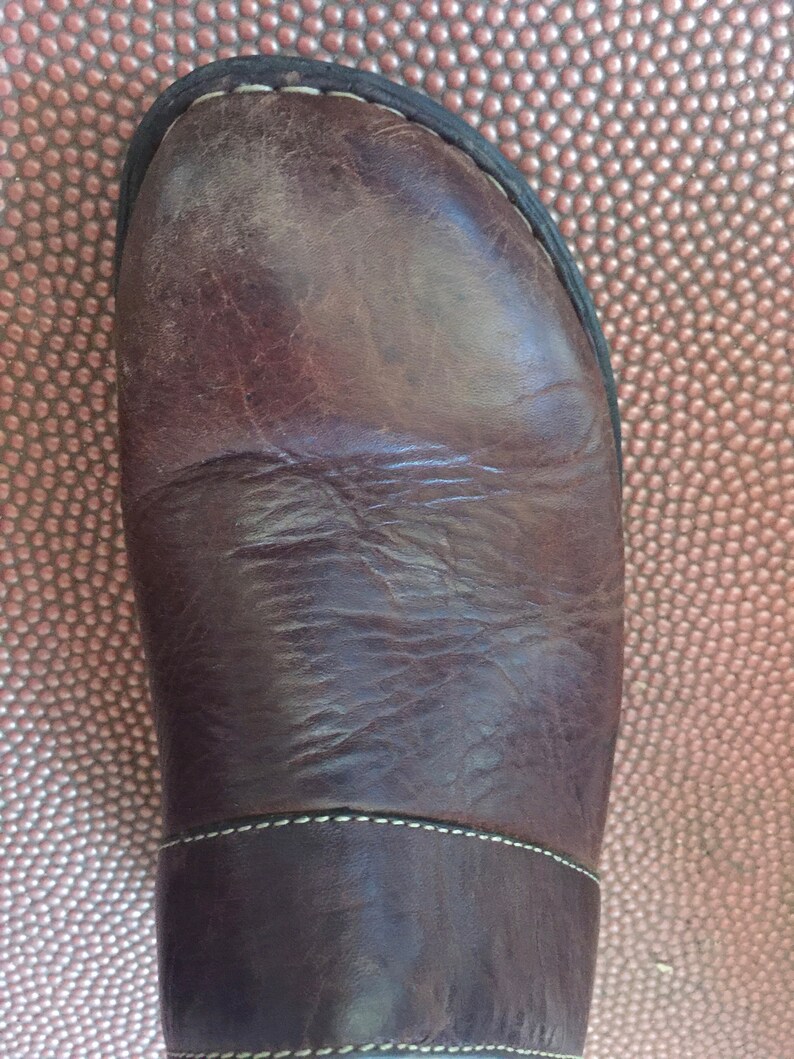 May include: Close-up of the top of a brown leather boot with a white stitch detail. The boot is on a brown textured surface.