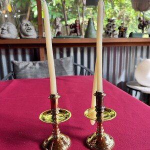 May include: Two gold candle holders with white candles on a red tablecloth.