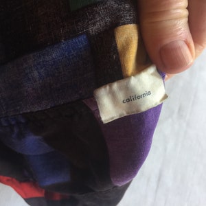 K&ouml;nnte beinhalten: Ein geometrisch gemustertes Hemd in Lila, Schwarz, Blau und Gelb mit einem wei&szlig;en Etikett, auf dem "california" steht.