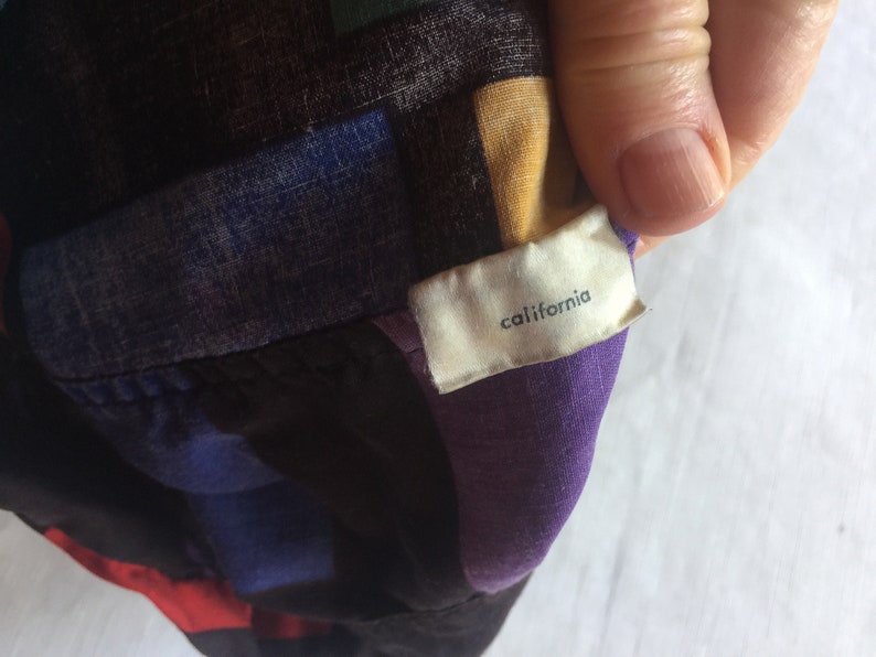 K&ouml;nnte beinhalten: Ein geometrisch gemustertes Hemd in Lila, Schwarz, Blau und Gelb mit einem wei&szlig;en Etikett, auf dem "california" steht.